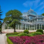 Vista do Palácio de Cristal em Porto cercado por jardins verdes e flores coloridas sob um céu azul claro.