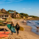 Praia de Punta del Diablo com surfistas, barcos de pesca na areia, casas à beira-mar e pessoas caminhando na praia sob céu azul.