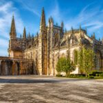 Mosteiro da Batalha com sua arquitetura gótica imponente, rodeado por árvores verdes e céu azul claro.