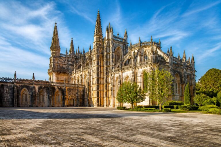 Mosteiro da Batalha com sua arquitetura gótica imponente, rodeado por árvores verdes e céu azul claro.