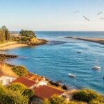 Vista da foz do rio Douro encontrando o oceano Atlântico com casas tradicionais, praia, pedras e barcos no mar sob um céu claro.