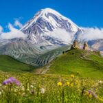 Montanhas nevadas de Kazbegi com uma igreja de pedra em um campo verde sob céu azul claro.
