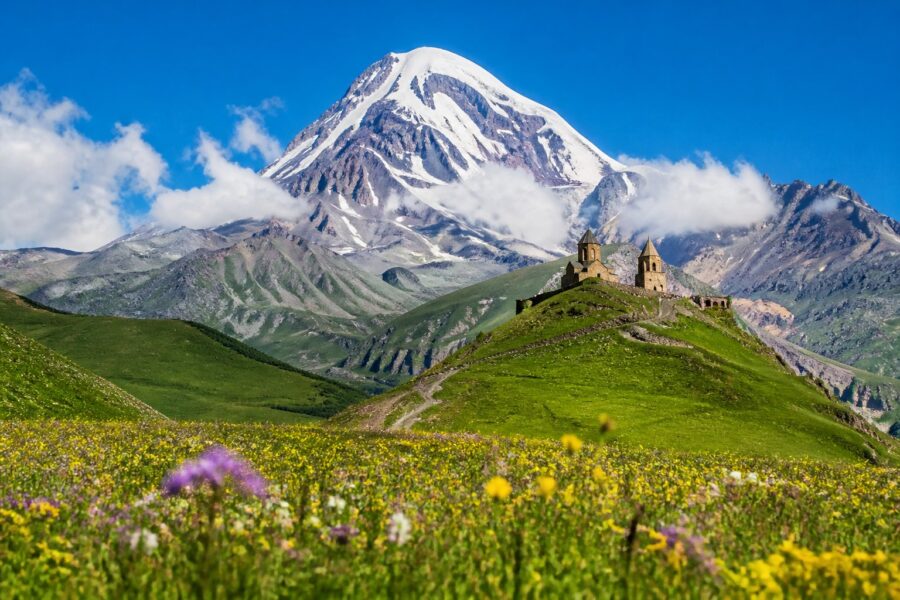 Montanhas nevadas de Kazbegi com uma igreja de pedra em um campo verde sob céu azul claro.