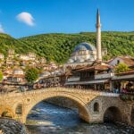 Vista da ponte histórica sobre o rio Bistrica em Prizren, com prédios tradicionais e a mesquita Sinan Pasha ao fundo, sob um céu azul claro.