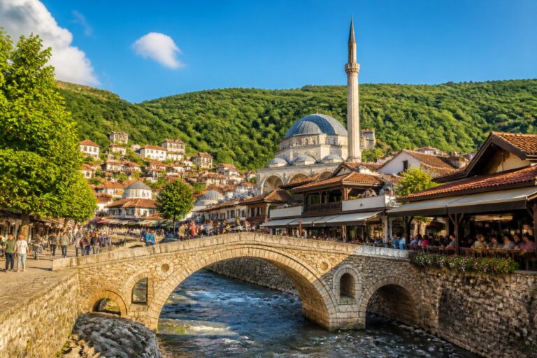Vista da ponte histórica sobre o rio Bistrica em Prizren, com prédios tradicionais e a mesquita Sinan Pasha ao fundo, sob um céu azul claro.