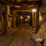 Túnel subterrâneo de uma mina de ouro com trilhos, suportes de madeira e rochas ao redor.