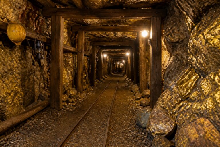 Túnel subterrâneo de uma mina de ouro com trilhos, suportes de madeira e rochas ao redor.