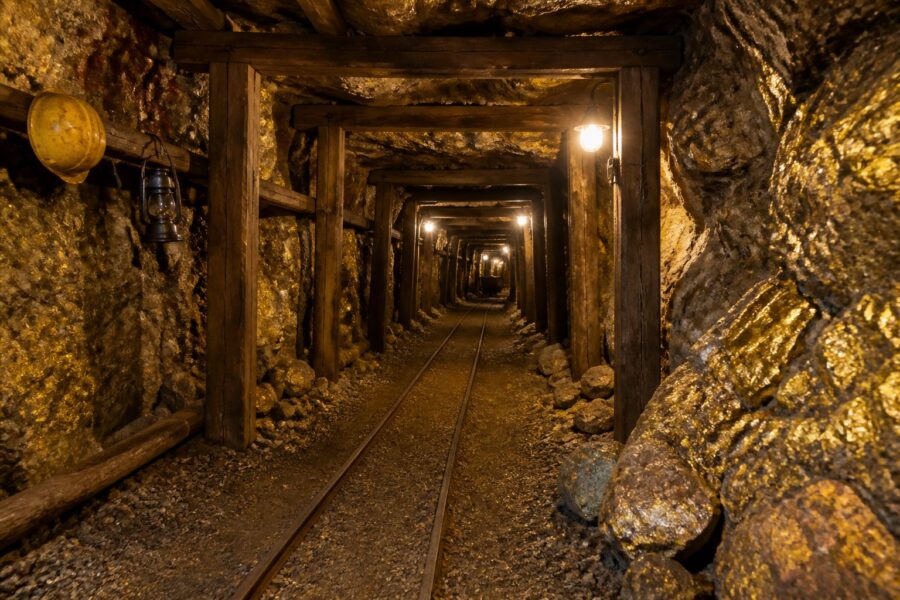 Túnel subterrâneo de uma mina de ouro com trilhos, suportes de madeira e rochas ao redor.