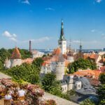 Vista da cidade de Tallinn com ruas de paralelepípedos, prédios históricos coloridos, torres de igrejas e pessoas em cafés ao ar livre em um dia ensolarado.