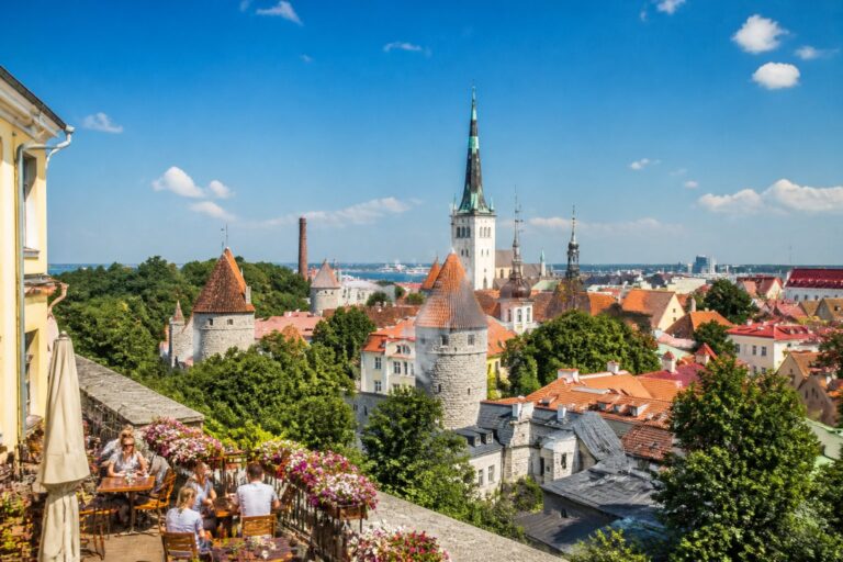 Vista da cidade de Tallinn com ruas de paralelepípedos, prédios históricos coloridos, torres de igrejas e pessoas em cafés ao ar livre em um dia ensolarado.