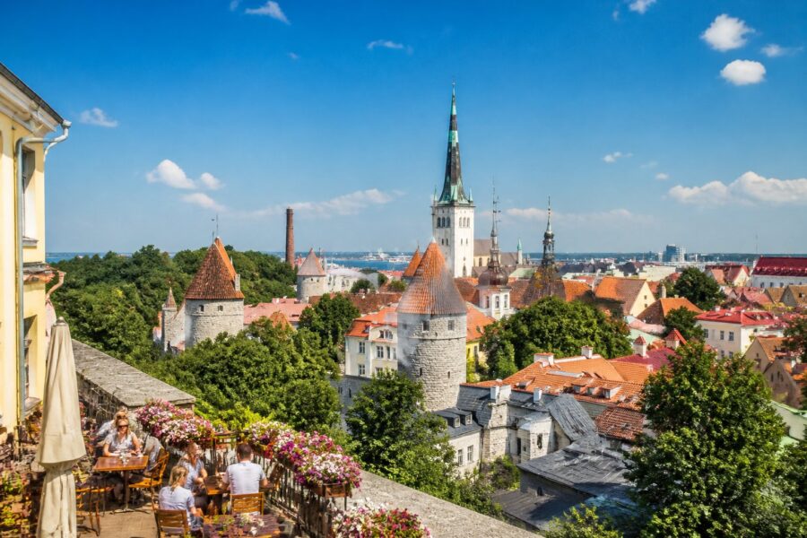 Vista da cidade de Tallinn com ruas de paralelepípedos, prédios históricos coloridos, torres de igrejas e pessoas em cafés ao ar livre em um dia ensolarado.