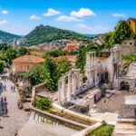 Teatro romano antigo em Plovdiv com casas coloridas ao redor e pessoas caminhando pelas ruas de pedra sob céu azul.